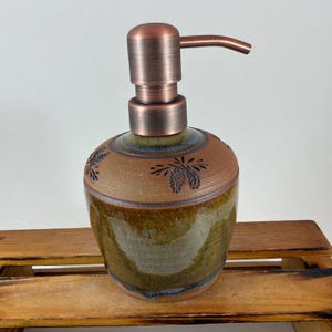 May include: A handcrafted ceramic soap dispenser with a copper-colored pump. The dispenser has a brown and green glazed finish with carved floral designs. The dispenser is a decorative and functional bathroom accessory.