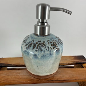 May include: A ceramic soap dispenser with a rounded base featuring a reactive glaze in shades of cream, light blue, and dark brown. The body has a textured, embossed pattern around the upper section and a brushed stainless steel pump mechanism.