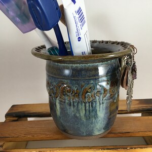 May include: A blue and brown ceramic utensil holder with a decorative design. The holder has a wide opening and is filled with a toothbrush, toothpaste, and earrings.