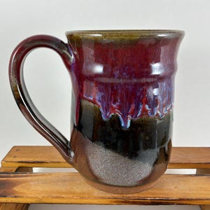 May include: A ceramic mug featuring a glossy glaze with a deep red and purple top section that transitions into a dark black and brown base. The mug has a large, curved handle and a smooth, reflective surface with visible drip patterns.