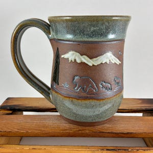 May include: A ceramic mug with a brown base and a green glazed rim and bottom section. The center features a relief design of white mountain peaks, a dark green pine tree, and three bears of varying sizes walking across the surface.