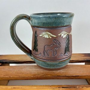 May include: A ceramic mug featuring a brown body with a blue-green glazed rim and base. The mug displays an embossed design of a moose standing between two pine trees, with two mountain peaks positioned above the moose in a light-colored glaze.