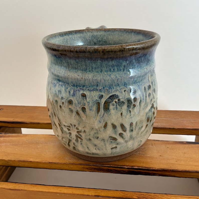 Pu&ograve; includere: Una tazza in ceramica fatta a mano con una superficie strutturata. La tazza presenta una sfumatura di colori, che passa da un bordo marrone scuro a sfumature di blu e bianco. La tazza ha un manico ed &egrave; adatta a bevande calde o fredde.