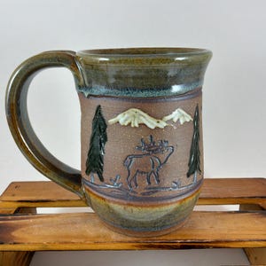 May include: A handcrafted ceramic mug with a nature-inspired design. The mug features a brown base with an embossed image of a moose, trees, and mountains. The rim and handle have a green and brown glaze. A unique piece for coffee or tea.