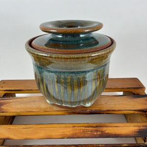 May include: A ceramic butter dish with a lid. The dish is a mix of brown, blue, and green hues. The lid has a circular handle. The dish is sitting on a wooden surface.