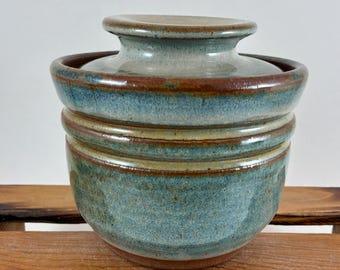 May include: A ceramic butter crock with a lid, featuring a mottled blue and brown glaze. The vessel has a rounded body with a horizontal ridge and a wide, flat-topped lid that fits securely into the top opening of the container.