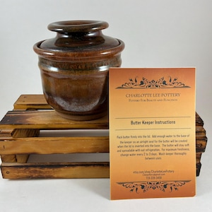 May include: A brown ceramic butter keeper with a lid, sitting on a wooden crate. The keeper has a rounded base and a bulbous lid. A card with "Butter Keeper Instructions" is next to the keeper. The card also has the text "Charlotte Lee Pottery".