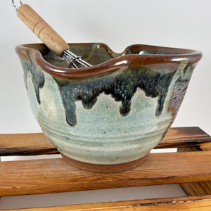 May include: A ceramic mixing bowl with a pour spout, featuring a light blue and cream base with dark brown and green drip glaze patterns. A metal whisk with a light-colored wooden handle rests inside the bowl, which has a wide, flared rim.
