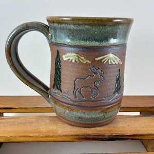 May include: A handcrafted ceramic mug with a brown base and a green rim. The mug features a detailed relief design of a moose, trees, and mountains. The handle is a dark green color. A rustic, nature-themed mug.