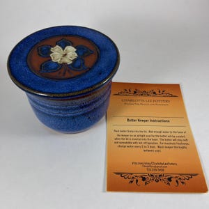 May include: A blue ceramic butter keeper with a brown circular center featuring a white flower design. Beside it is an orange instruction card titled Butter Keeper Instructions from Charlotte Lee Pottery with text detailing usage and care.