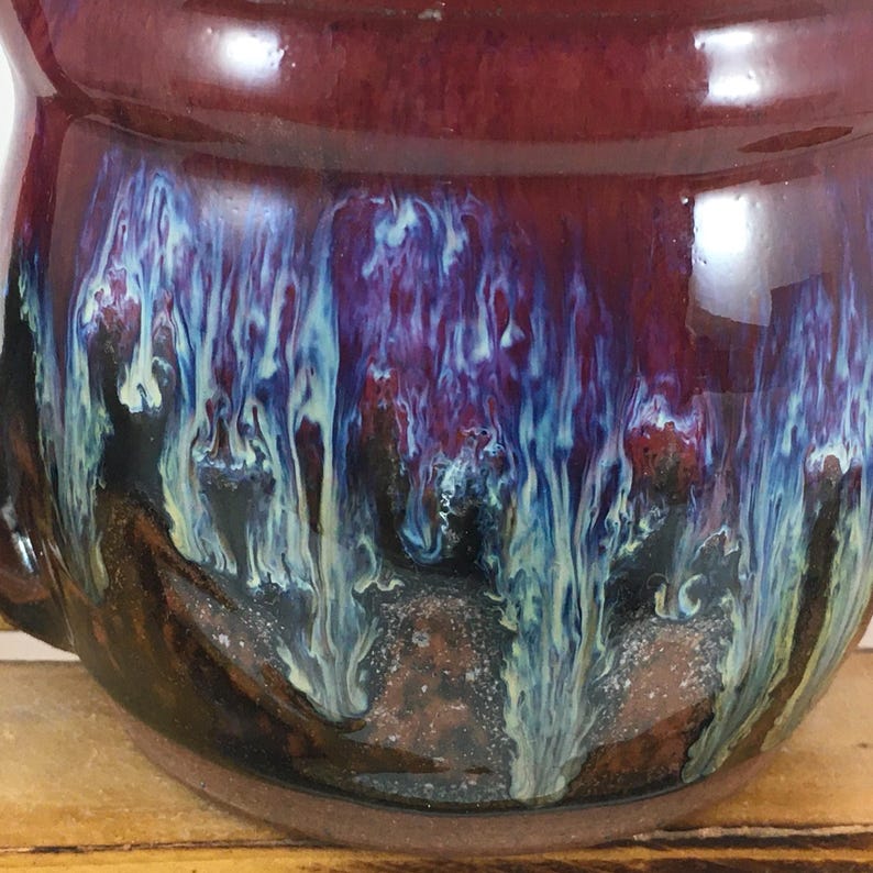 以下が含まれることがあります： 光沢仕上げの手作りセラミックマグのクローズアップ。マグカップは、深いバーガンディ色の外観から、青、紫、クリーム色の渦巻きへと変化します。内側は濃い茶色です。温かい飲み物を楽しむためのユニークな作品です。
