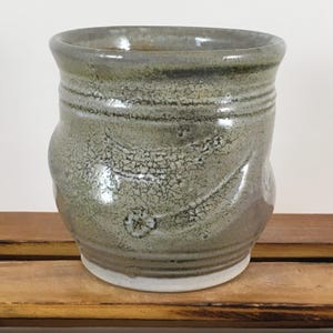 May include: A handmade ceramic mug with a textured, crackled surface in shades of gray and green. The mug has a wide, slightly flared rim and a white base. The design includes horizontal lines and a subtle floral motif.