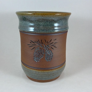 May include: A handmade ceramic tumbler with a brown body and a carved pinecone design. The rim and base feature a green-blue glaze, and the top rim has a brown glaze. This rustic mug is suitable for beverages.