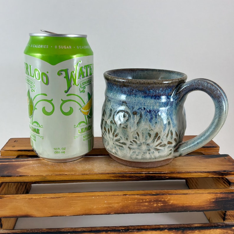 Pu&ograve; includere: Una tazza in ceramica fatta a mano con smalto blu e marrone, con un motivo floreale. La tazza &egrave; accanto a una lattina di acqua frizzante al gusto lime. La lattina &egrave; verde e bianca con la scritta "WATERLOO" e "LIME".