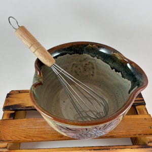 May include: A ceramic mixing bowl with a speckled beige interior and a dark brown rim, featuring a decorative floral pattern on the exterior. A metal whisk with a light-colored wooden handle rests inside the bowl, which has a pouring spout.