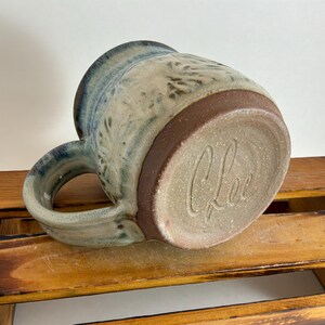 Pu&ograve; includere: Tazza in ceramica fatta a mano con smalto blu e beige. La tazza presenta una base e un manico marroni, con la firma dell'artista sul fondo. La tazza &egrave; progettata per bevande calde.