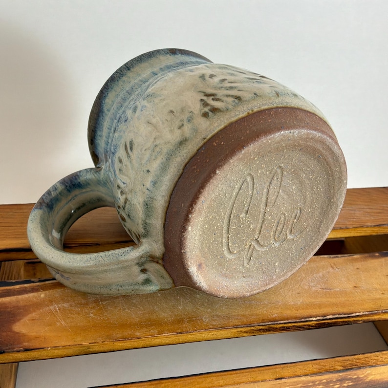 Pu&ograve; includere: Tazza in ceramica fatta a mano con smalto blu e beige. La tazza presenta una base e un manico marroni, con la firma dell'artista sul fondo. La tazza &egrave; progettata per bevande calde.