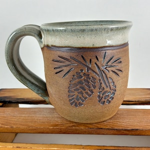May include: A ceramic mug with a brown, textured body and a smooth, light green glazed rim. The front features an embossed design of two pinecones with needles. The handle is thick and curved, matching the green glaze of the upper rim.