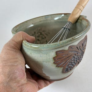 May include: A ceramic mixing bowl with a light blue and cream glaze, featuring a textured pinecone relief on the exterior and a pouring spout. A metal whisk with a light-colored wooden handle is placed inside the bowl, which has a smooth interior.