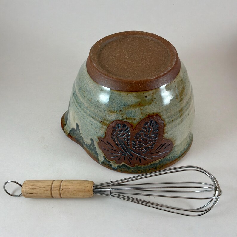 May include: A ceramic mixing bowl with a speckled light green and tan glaze, featuring a dark brown embossed pinecone design on the side, paired with a metal wire whisk that has a light-colored wooden handle and a small metal hanging loop.