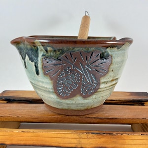 May include: A ceramic mixing bowl with a light green and brown glaze featuring a dark brown embossed pinecone design on the front. The bowl has a pouring spout and a wooden pestle handle extending from the top, showcasing a rustic, earthy finish.