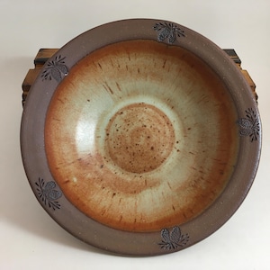 May include: A handmade ceramic bowl with a wide rim and a shallow interior. The bowl features a warm color palette of brown, tan, and cream, with a textured surface. Decorative pinecone designs are carved around the rim.