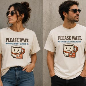 Puede incluir: Una camiseta de manga corta color crema con el texto "PLEASE WAIT. MY COFFEE HASN'T CLOCKED IN." sobre un gráfico de una taza de café sonriente que lleva una correa naranja con una tarjeta de identificación "STAFF".