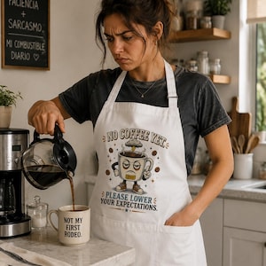 May include: A white apron featuring a graphic of a grumpy coffee mug with a storm cloud above it, surrounded by coffee beans. The text reads NO COFFEE YET, PLEASE LOWER YOUR EXPECTATIONS. The apron includes a front pocket and adjustable neck strap.