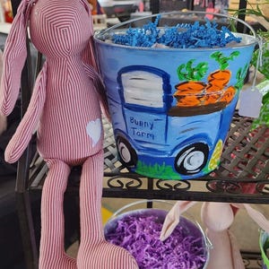 May include: A pink and white striped bunny plush toy with long ears and a white heart-shaped patch. Beside it is a blue bucket with a painted image of a truck carrying carrots, with the words "Bunny Farm". The bucket is filled with blue paper shreds.