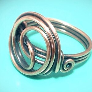 May include: A copper-colored wire ring featuring a multi-layered spiral design. The metal wire is coiled into a circular pattern that forms the centerpiece of the ring, with the wire continuing to create the band that wraps around the finger.