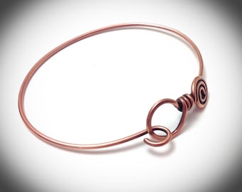 May include: A copper bangle bracelet with a spiral wire detail. The bracelet is open and has a simple, minimalist design.