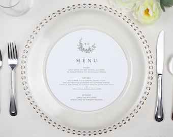 May include: A white menu card with a black border and gold accents. The menu card is on a white plate with a gold beaded rim. The menu card has the text "EI" in a black circle with a floral design. The menu card has the text "MENU" in black. The menu card has the text "SALAD", "MARKET GREENS WITH BABY TOMATOES, CUCUMBERS, AND RADISHES DRESSED WITH WHITE BALSAMIC DRESSING", "ENTREE", "GRILLED PETITE FILET AND PRETZEL CRUSTED CHICKEN WITH STOUT REDUCTION ACCOMPANIED WITH DAUPHINOISE POTATOES, ASPARAGUS, BABY CARROTS AND A SPINACH ARTICHOKE STUFFED ROMA TOMATO", "CHEF'S CHOICE SEASONAL VEGAN ENTREE", "DESSERT", "MARBLE CAKE WITH LAYERED BAVARIAN CREAM AND RASPBERRY PRESERVES".