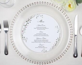 May include: A white plate with a gold rim and a floral design menu card on top. The menu card lists the salad course, entree, vegetarian option, dessert, and wedding cake. The menu card has a floral border and a handwritten font.