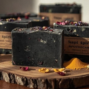 May include: Four dark charcoal-colored soap bars with floral and botanical accents are displayed on a wooden slab. The soaps have a rustic appearance and are labeled "Hapi Spirit Apothecary". A small pile of yellow powder and rose petals are scattered around the soaps.