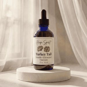 May include: A dark blue glass bottle with a dropper, labeled "Hapi Spirit Turkey Tail Double-Extracted Tincture, 2 fl oz / 60 ml." The bottle sits on a small, round, white pedestal against a soft, neutral background.