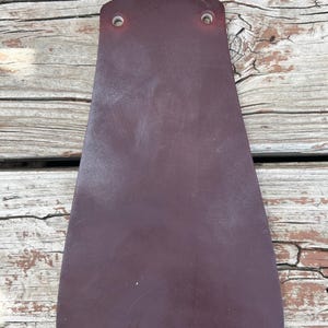 Può includere: Un pezzo di pelle bordeaux, con una base arrotondata e due fori nella parte superiore. La pelle ha una consistenza liscia e una leggera lucentezza. Sembra far parte di un prodotto più grande.