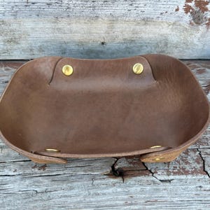 以下が含まれることがあります： 真鍮のリベットが付いた茶色の革製のトレイ。トレイは角が丸みを帯びた長方形で、側面にわずかなカーブがあります。