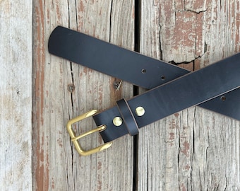 May include: A black leather belt with a brass buckle and two brass rivets. The belt is crossed over itself and laid on a wooden surface.