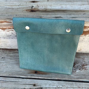 Op de afbeelding: Een teal leren etui met een klep die is vastgezet met twee zilveren drukknopen. De etui heeft witte stiksels langs de randen en een getextureerd oppervlak. De achtergrond is verweerd hout.