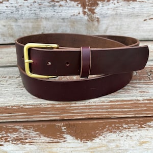 May include: A dark brown leather belt with a brass buckle. The belt is rolled up and lying on a wooden surface.