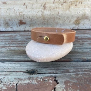May include: A light brown leather bracelet with a gold-colored button clasp. The bracelet is resting on a smooth, white stone, set against a weathered wooden background. The bracelet appears to be approximately 0.75 inches wide.