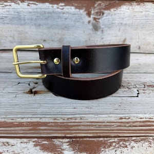 May include: A dark brown leather belt featuring a polished gold-tone metal buckle and two matching gold-tone metal rivets securing the belt loop. The belt has a smooth finish with visible edges and a classic rectangular buckle frame design.