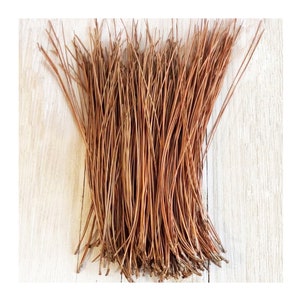 May include: A bundle of thin, brown pine needles, arranged in a fan shape on a light wood surface.