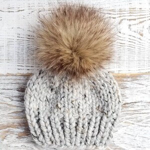 May include: A gray knitted beanie with a brown faux fur pom pom.