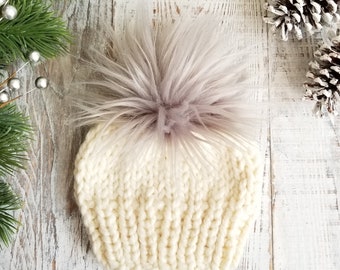 May include: A white knitted beanie with a fluffy gray pom pom. The beanie is made of a soft, chunky yarn and has a slouchy fit.