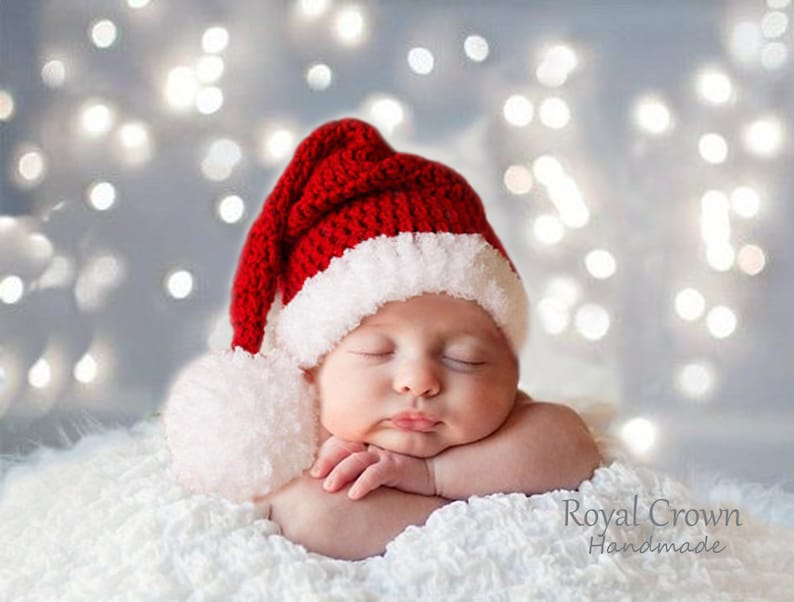 Pu&ograve; includere: Un bambino che dorme con un cappello di Babbo Natale rosso e bianco a maglia con un pon pon bianco. Il bambino &egrave; sdraiato su una coperta bianca e soffice con uno sfondo sfocato di luci bianche.