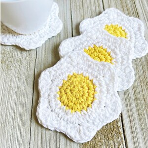 May include: Four crocheted white star-shaped coasters with yellow centers, resembling fried eggs. The coasters are arranged in a stack on a wooden surface.