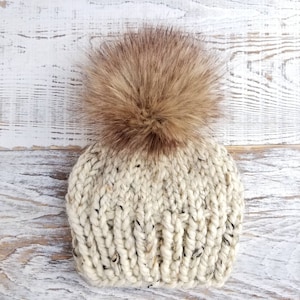 May include: A white knitted beanie with a brown faux fur pom pom. The beanie is made with a chunky yarn and has a slouchy fit.