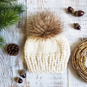 May include: A white knitted beanie with a brown faux fur pom pom. The beanie is made of a soft, chunky yarn and has a ribbed texture.