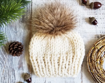 May include: A white knitted beanie with a brown faux fur pom pom. The beanie is made of a soft, chunky yarn and has a ribbed texture.
